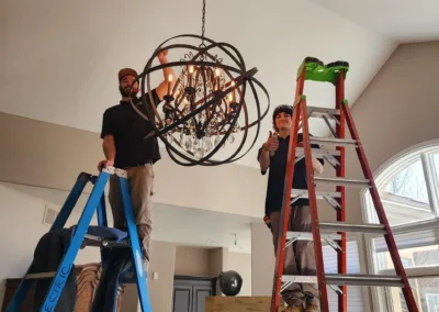 Two men working on a chandelier in a living room.